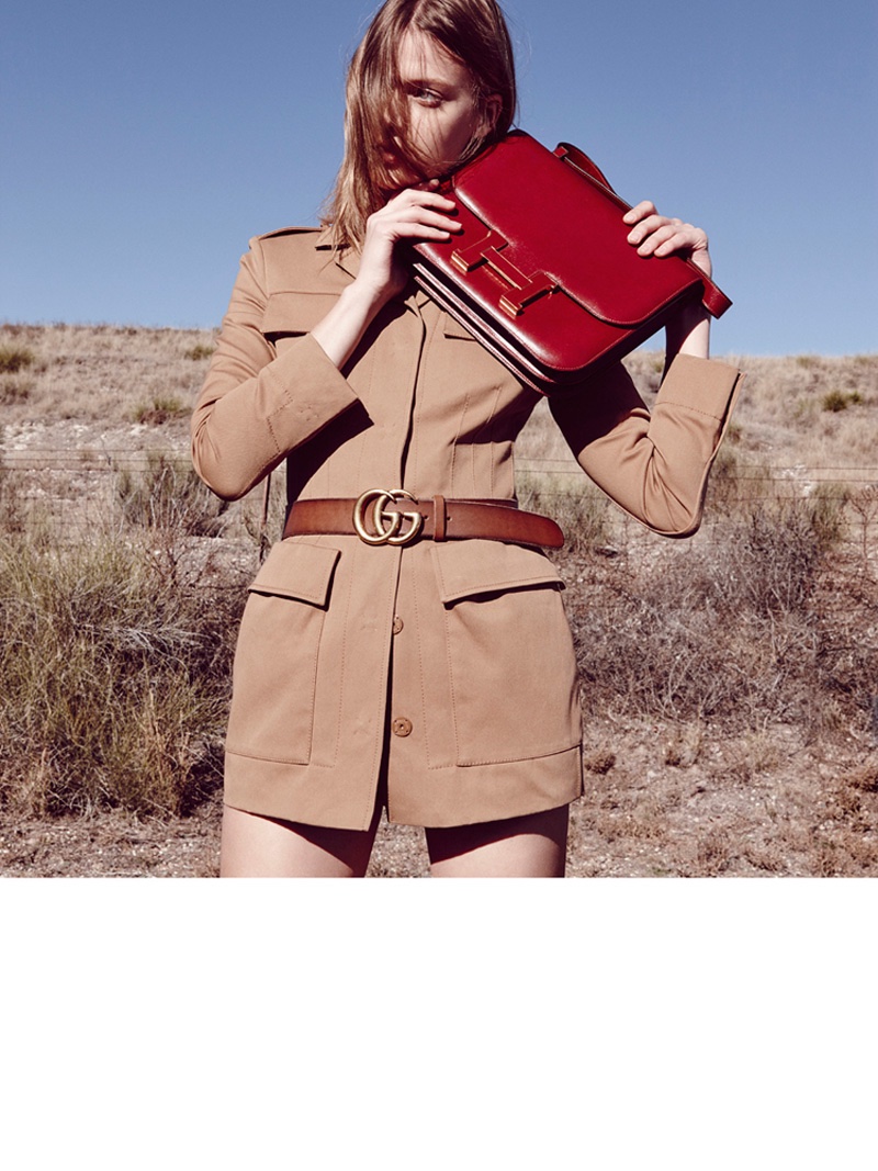 Tess poses in a brown jacket featuring a Gucci belt and oversized red Hermes clutch