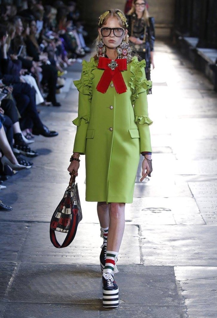 A model walks the runway at Gucci’s resort 2017 show wearing a green coat with ruffle-embellished sleeves and platform sneakers
