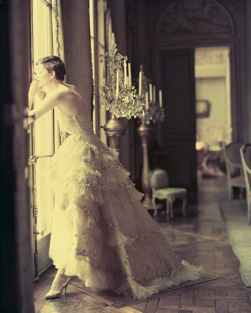 ca. May 1950 --- A fashion model peers out the window of an elegant home wearing a white Christian Dior evening gown decorated with tiers of ruffles. --- Image by © Norman Parkinson/Corbis