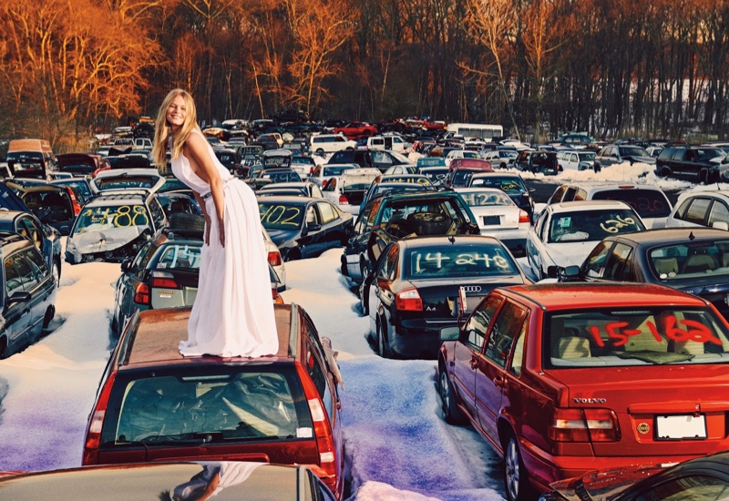 Modeling in a junkyard, Anna Ewers smiles in a Carolina Herrera maxi dress with pleats and a belt