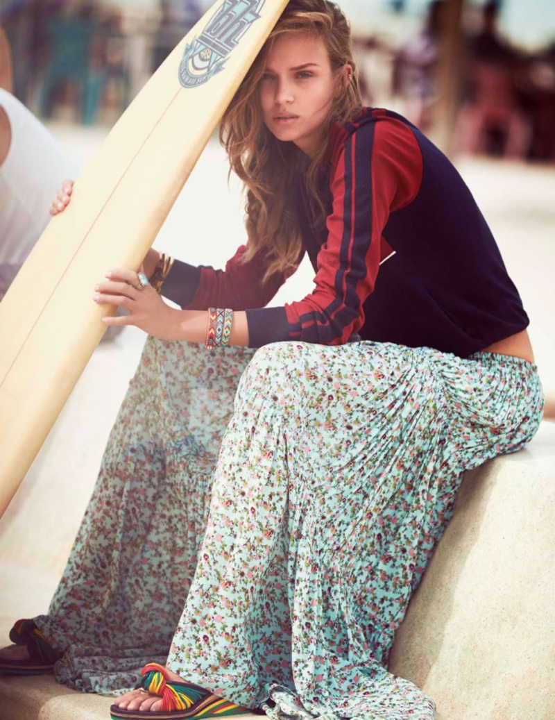Holding a surfboard, Josephine models a tracksuit jacket with a pleated maxi skirt and open-toe sandals
