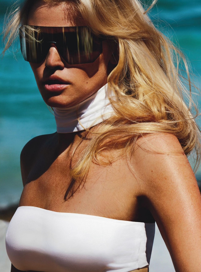 Photographed on the beach, Lara Stone models a white swimsuit with black visor sunglasses