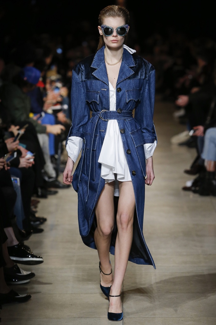 A model walks the runway at Miu Miu's fall-winter 2016 show wearing a denim coat belted at the waist with a white dress