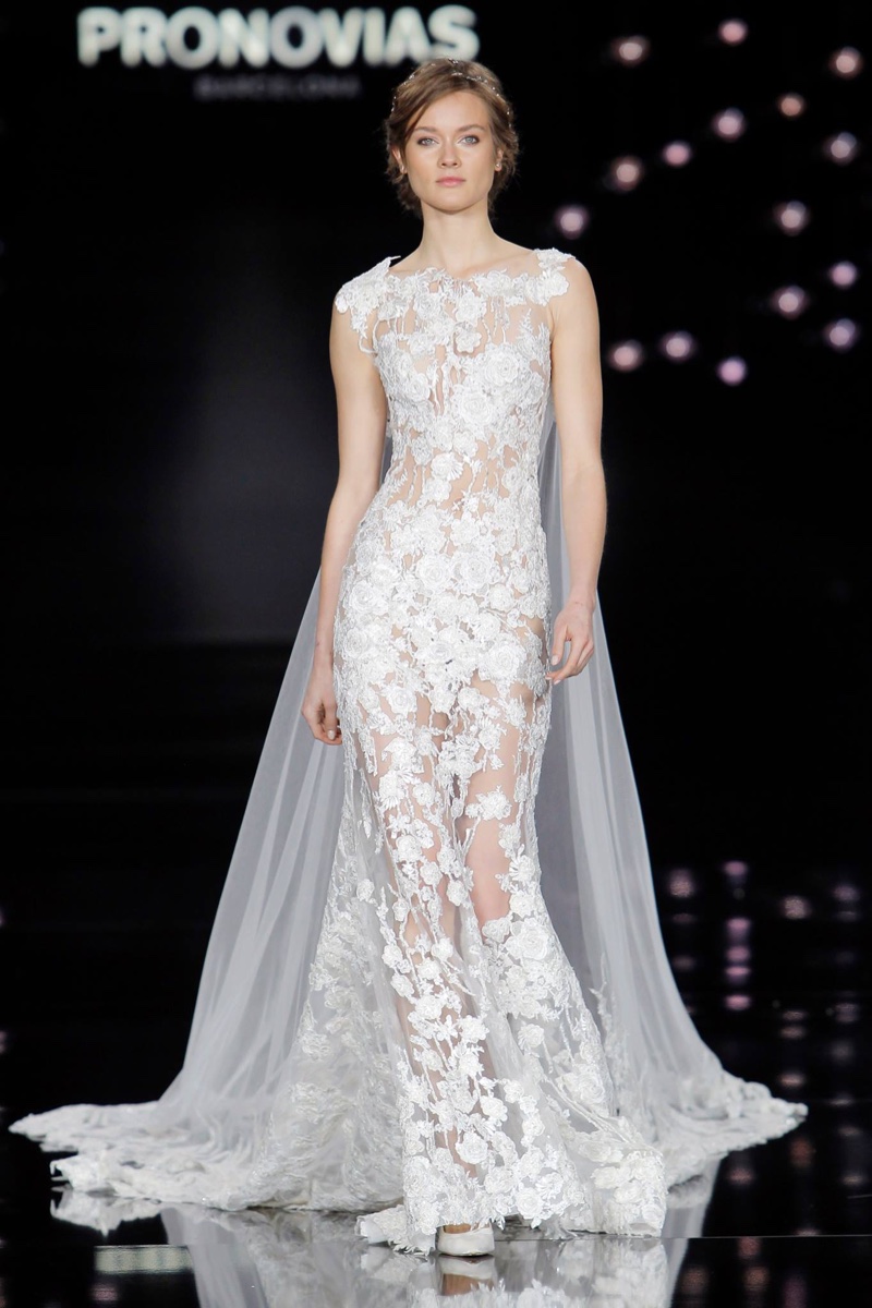 Jac Jagaciak walks the runway at Atelier Pronovias 2017 show wearing a sleeveless wedding dress with floral embellishments and a sheer cape