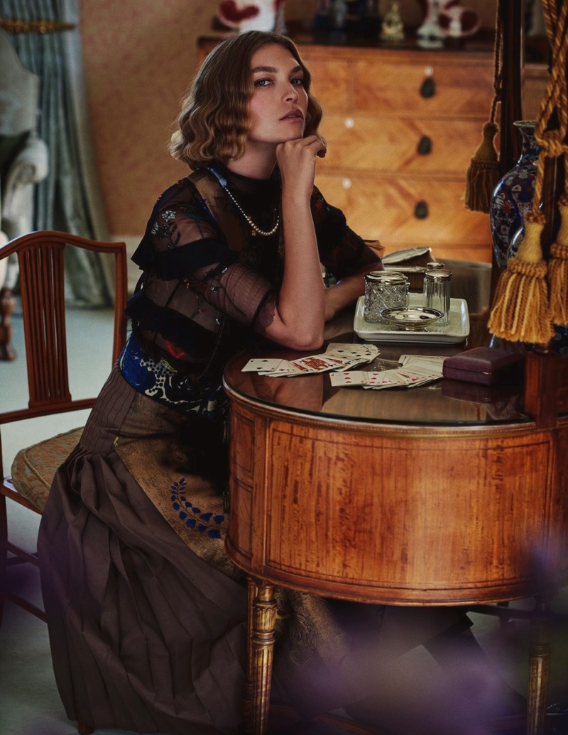 Sitting at a desk, Arizona Muse wears embellished top and pleated skirt