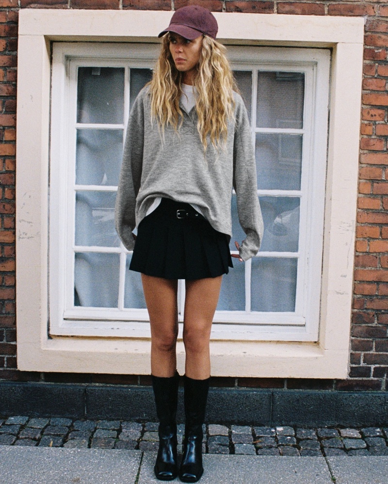 Olivia Vinten poses in baseball hat, polo sweatshirt, pleated skort, and ankle boots from Zara.