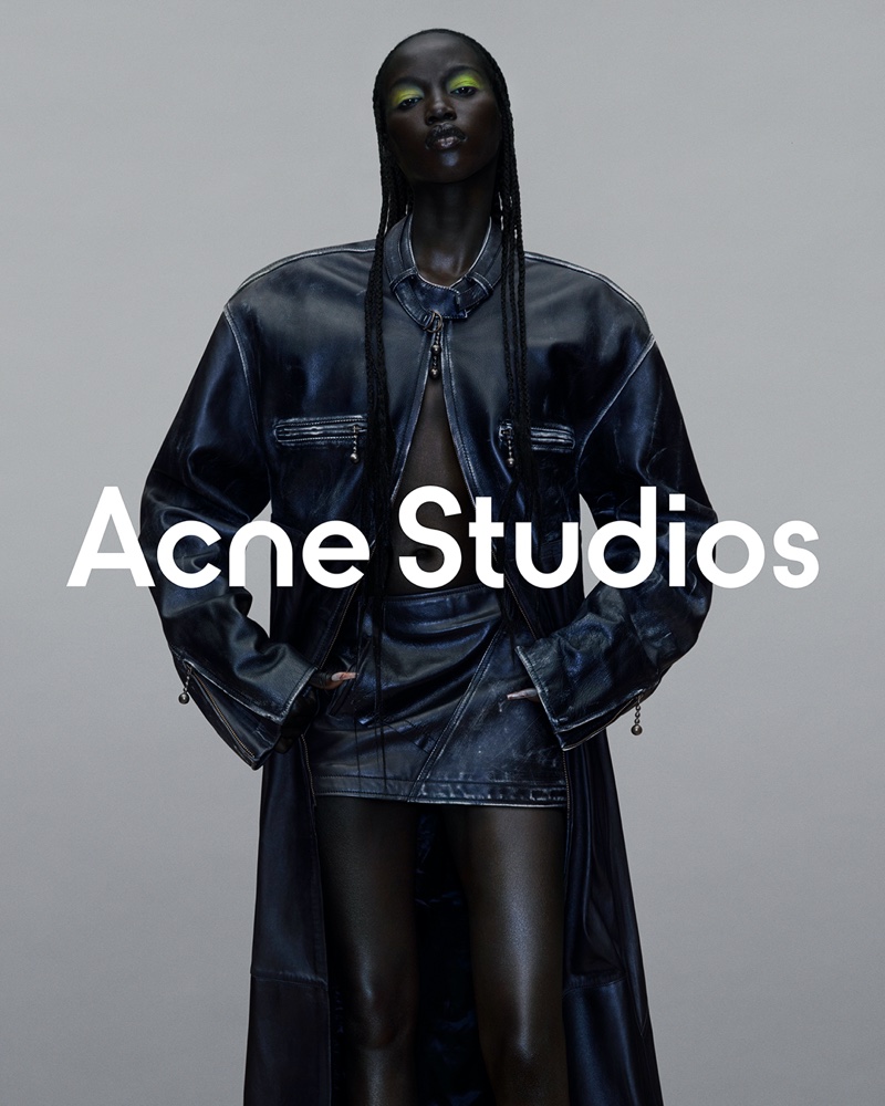 Model Anok Yai shows a glossy coat and mini skirt ensemble with striking neon eye makeup for Acne Studios' Fall 2023 campaign.