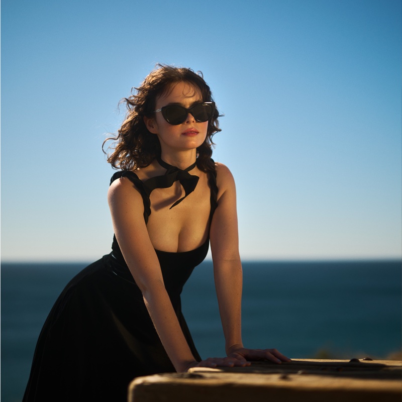 Overlooking the serene ocean, Micaela Wittman strikes a poised stance in a chic black dress with a bold neckline.