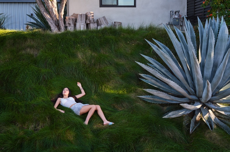 Singer Olivia Rodrigo lounges on a lush, grassy hillside, effortlessly blending with the natural landscape in a casual, sun-drenched setting.