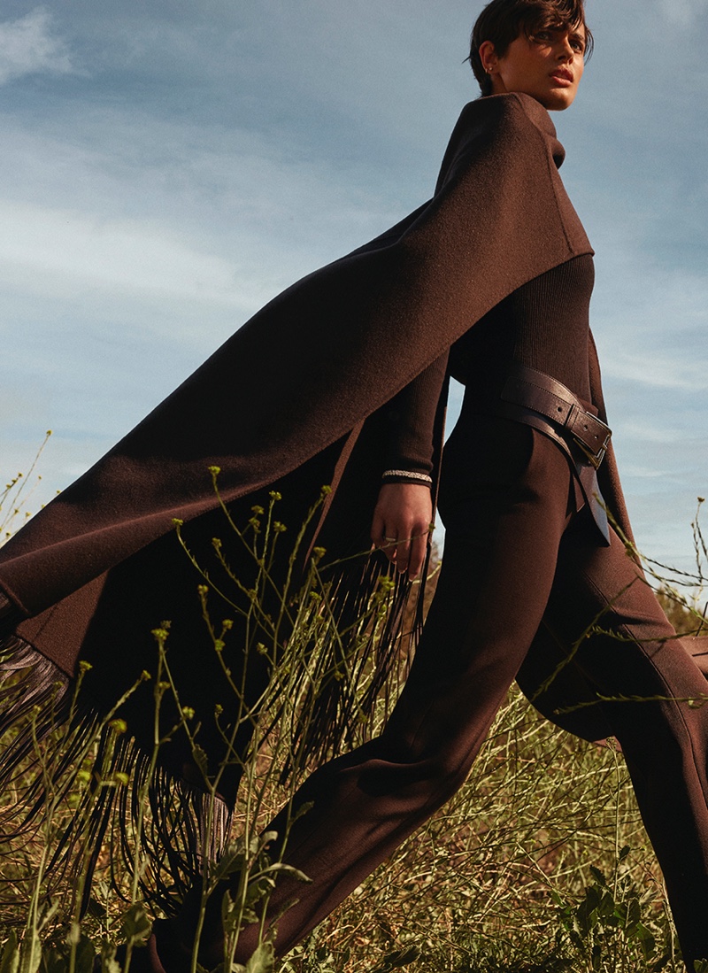 Taylor Hill strikes a dynamic pose in a flowing dark cape against a serene field, embodying elegance in motion.