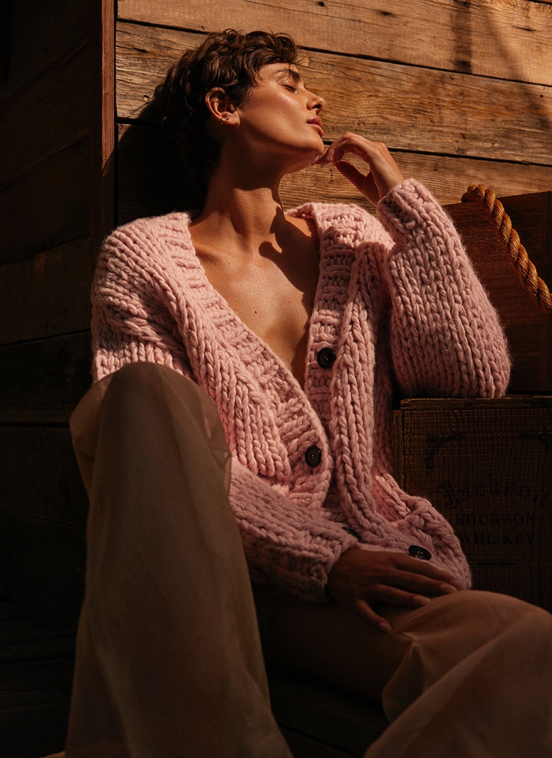 Bathed in golden sunlight, Taylor Hill reflects a moment of tranquility, clad in a plush pink cardigan, against a rustic wooden backdrop.