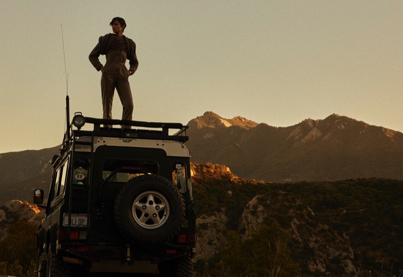 Commanding the scene, Taylor Hill stands atop a rugged vehicle, her silhouette set against the backdrop of a majestic mountain range.