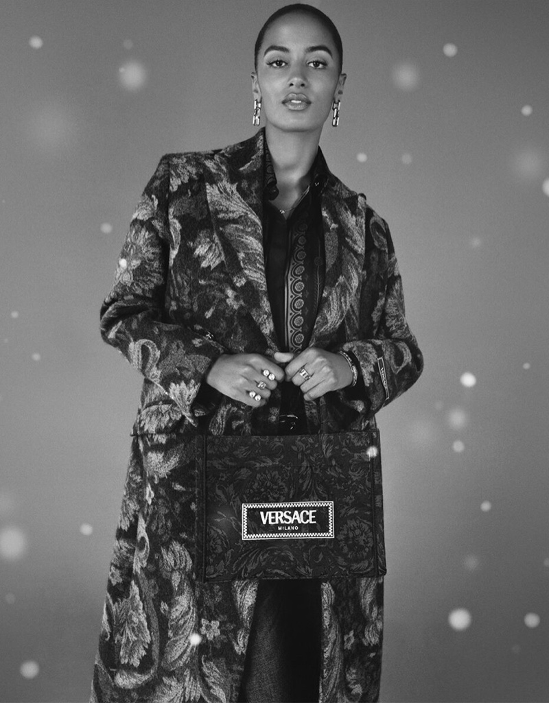Sacha Quenby poses under a gentle snowfall wearing a Barocco print coat and tote bag for Versace's holiday 2023 collection. 
