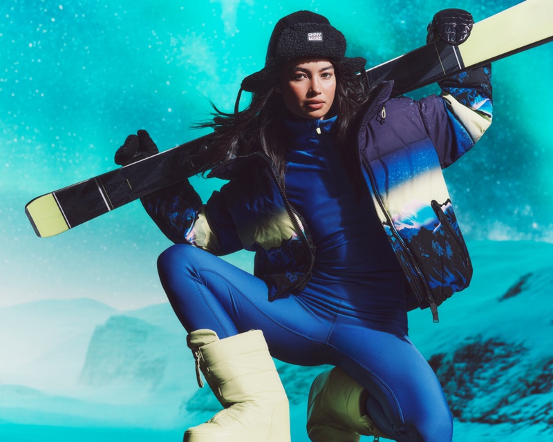 Kelsey Merritt carries her snowboard, dressed in a blue DKNY Tech ski jumpsuit and puffer jacket with a Northern Lights inspired print.
