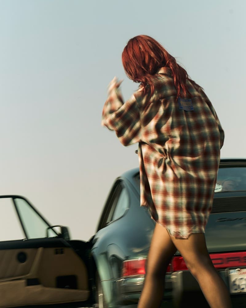 Dua Lipa, captured mid-movement, her auburn hair flowing, turns away from the camera, her plaid shirt in the spotlight next to a Porsche car.