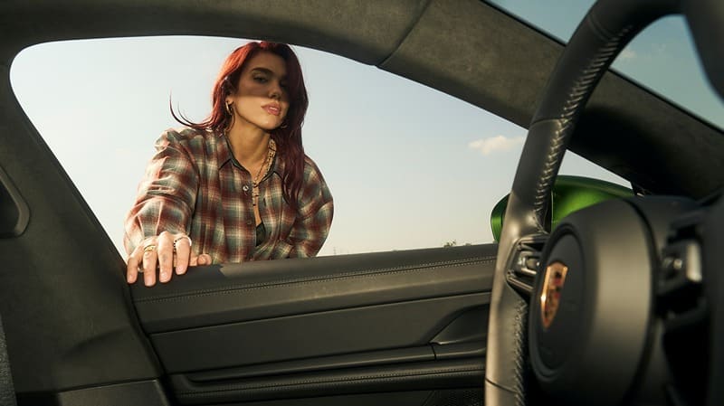 Dua Lipa leans on the door of the Porsche, with the sleek interior framing the shot, hinting at an adventure waiting to unfold on the road.