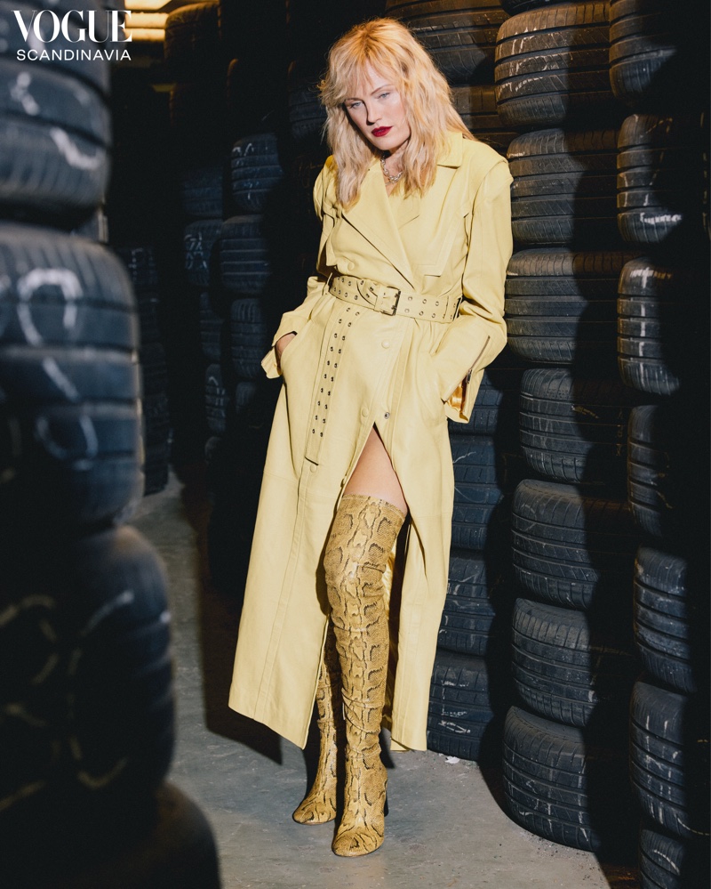 Malin Åkerman stands out in a vibrant yellow trench and leather boots against the backdrop of a garage setting.