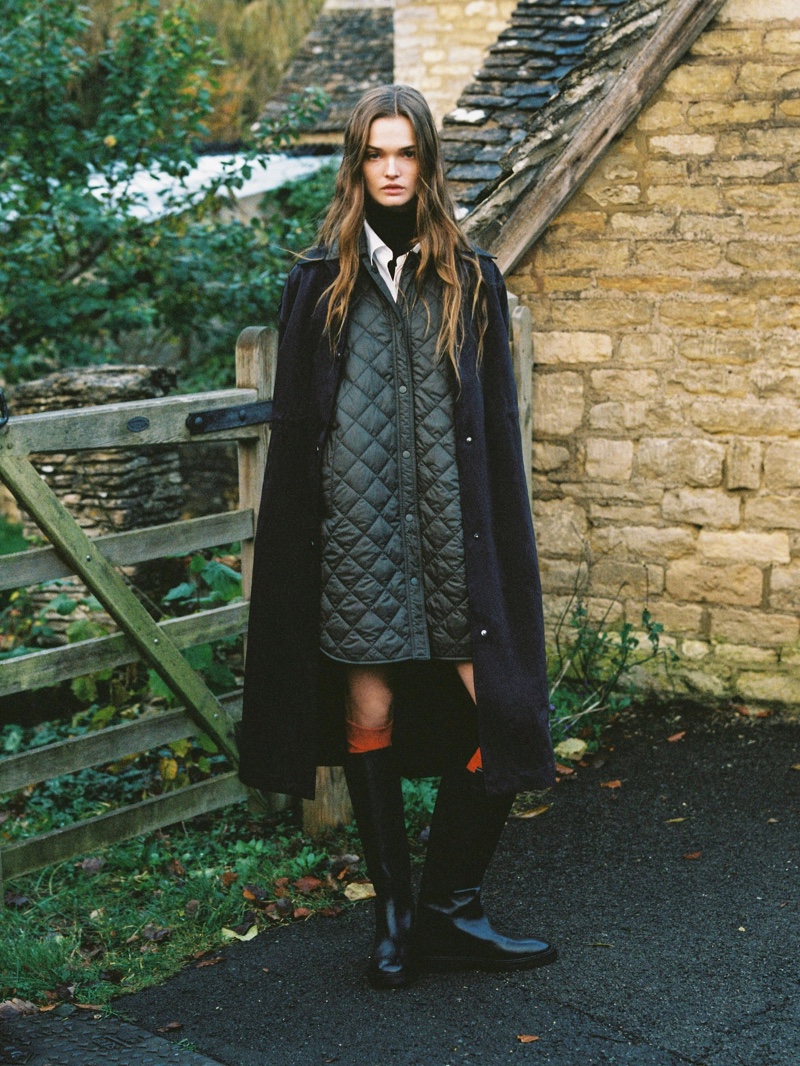 Massimo Dutti features a winter ensemble with a quilted parka and long coat, accentuated by a pop of orange against the rustic backdrop of an English countryside gate.
