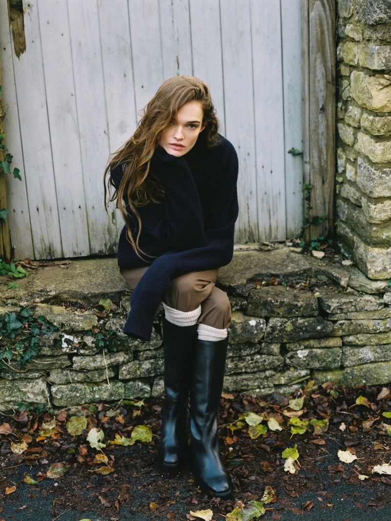 Lulu Tenney sits contemplatively in a cozy navy Massimo Dutti sweater and taupe trousers, complemented by knee-high boots, amidst a quaint village setting.