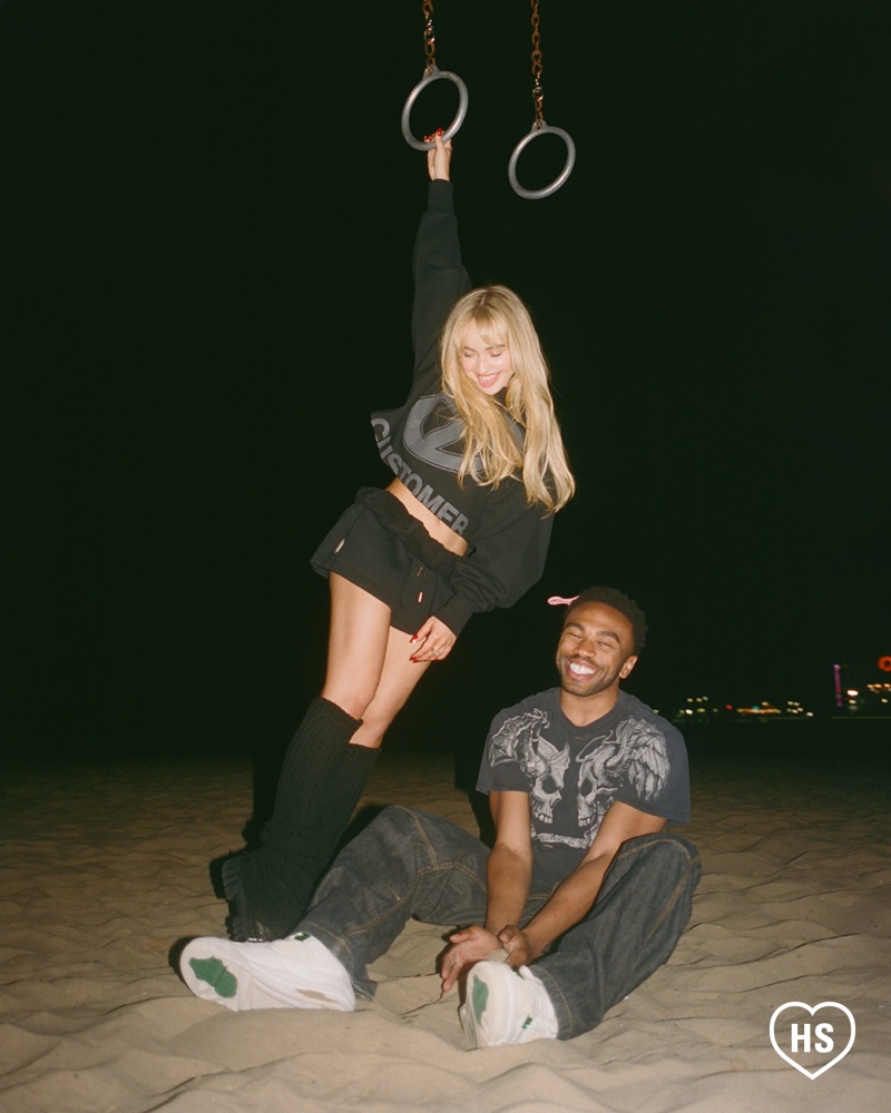 On a beach, Sabrina swings from rings as Kevin Abstract supports her, showcasing a spirited and carefree moment.