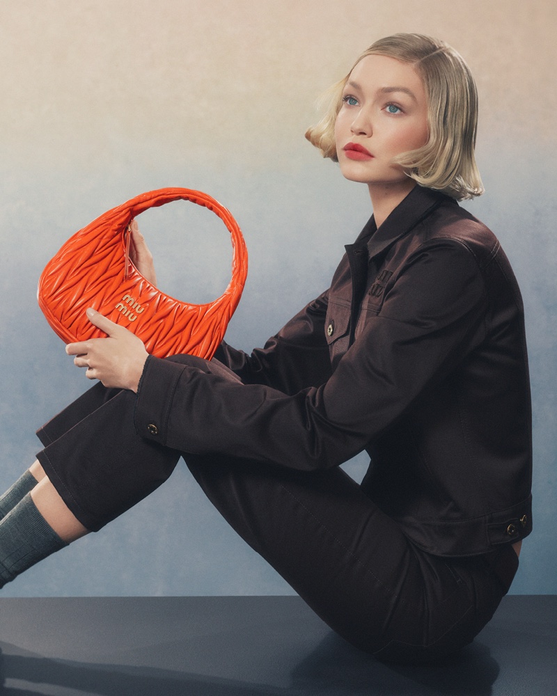 Embracing an orange style, Gigi Hadid showcases Miu Miu's handbag in a chic chocolate jacket for 2024 ad.